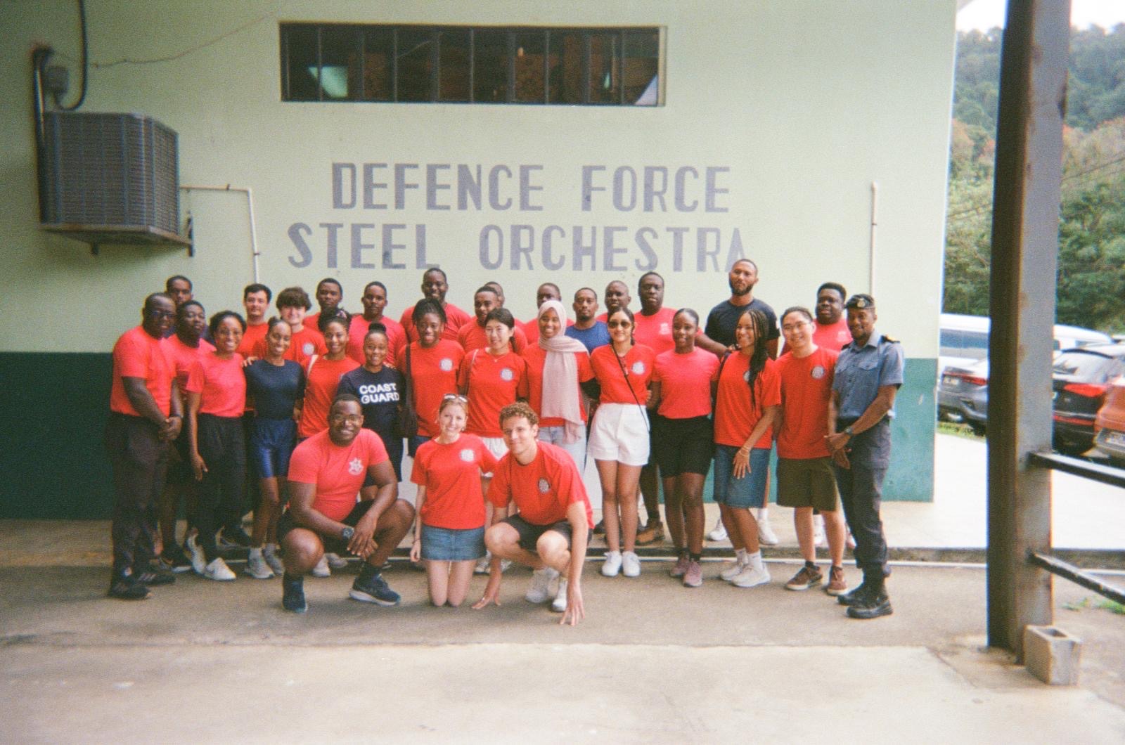 The ensemble standing with members of the Defence Force Steel Orchestra outside their panyard, in front of a wall painted with the orchestra's name.