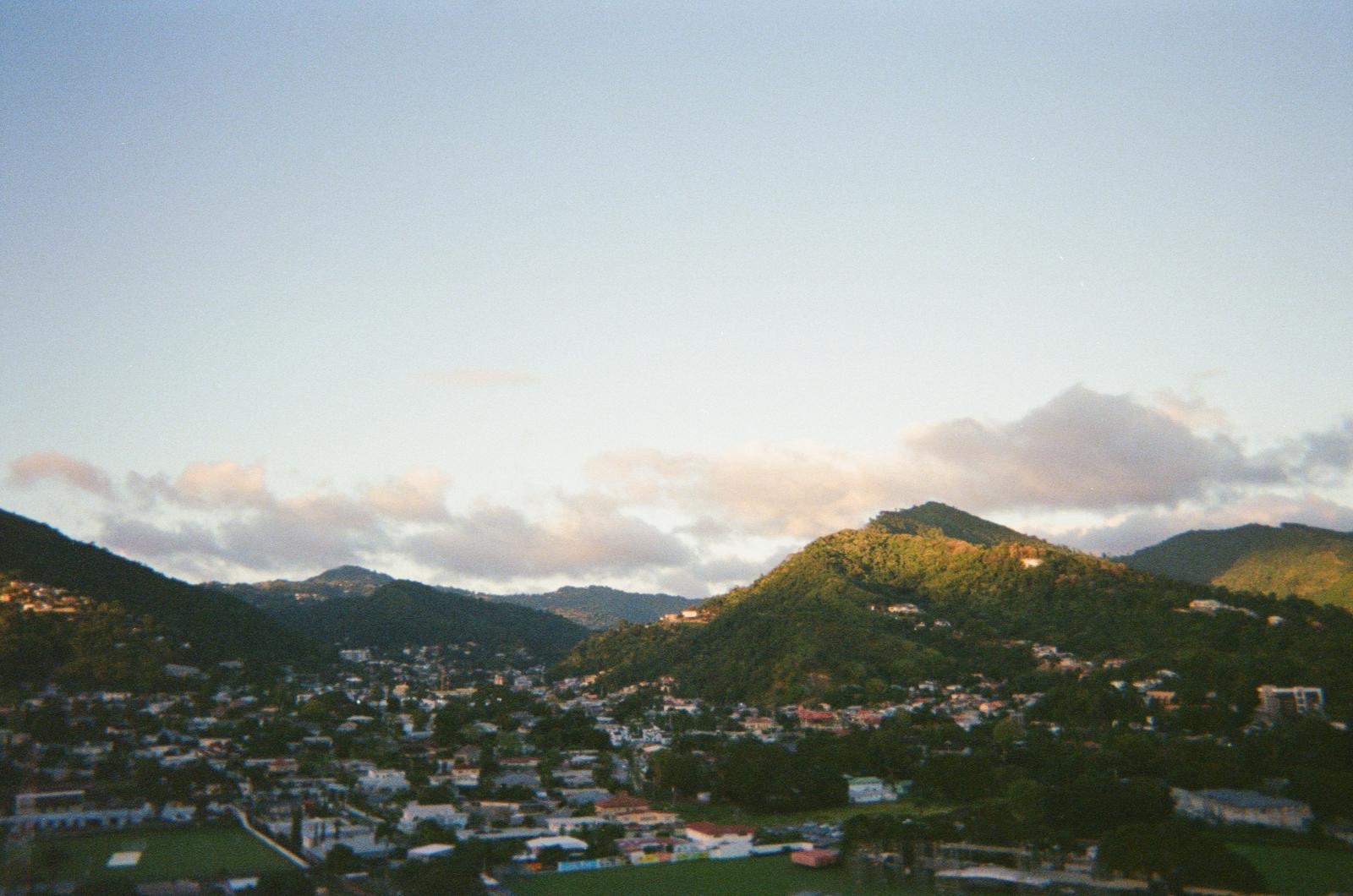 The hills above Port of Spain at golden hour, houses scattered across the green slopes under a wide sky.