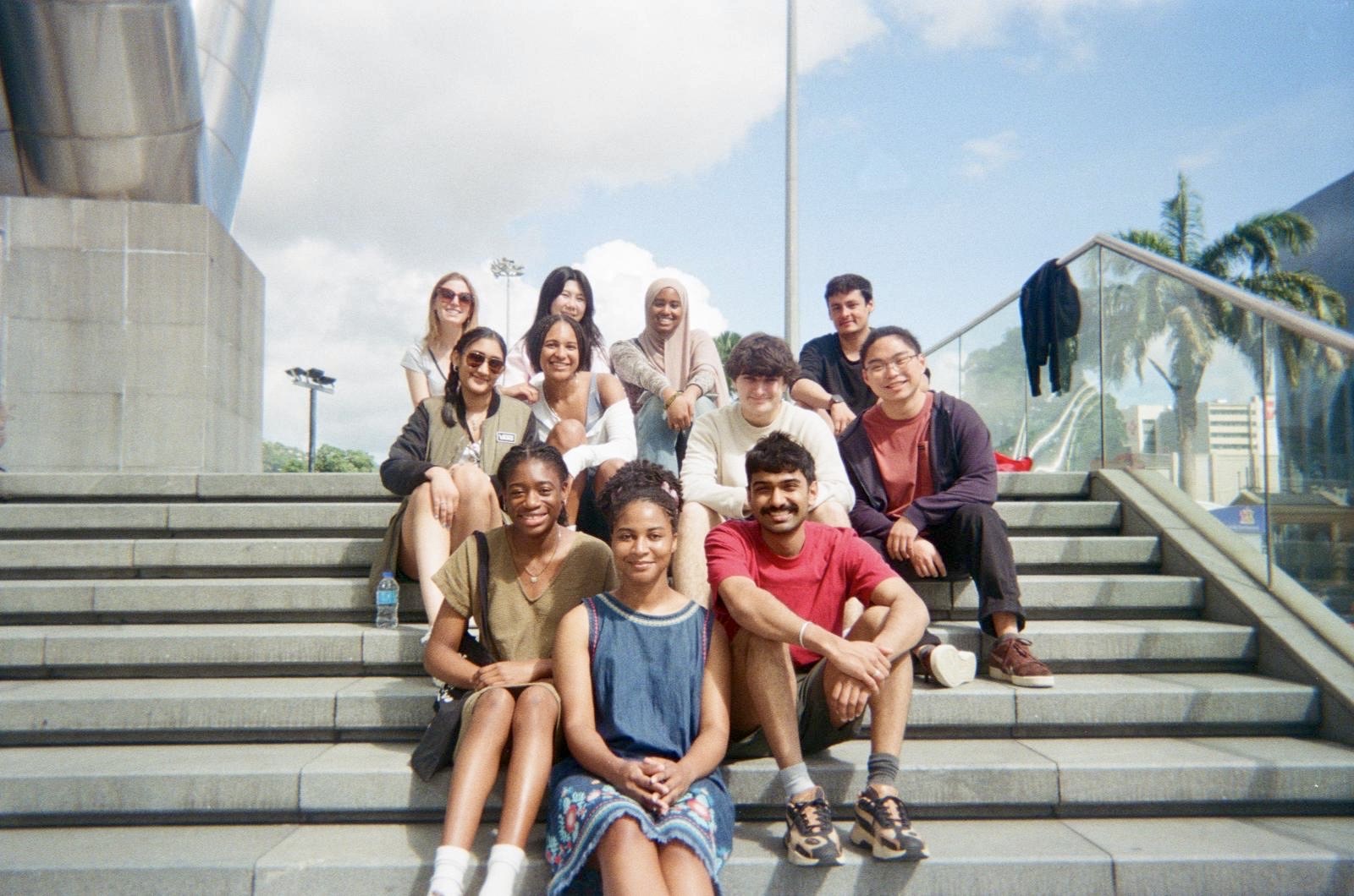 The ensemble sitting together on the steps outside the National Academy for the Performing Arts in Port of Spain, under a bright sky.