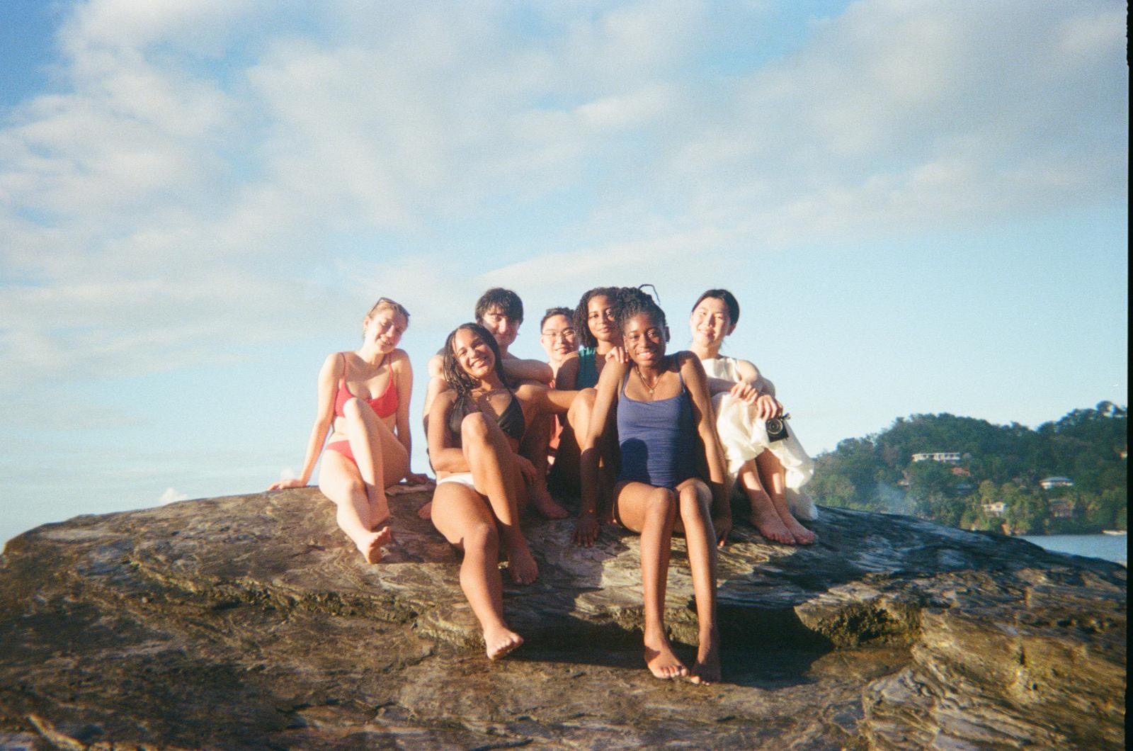 Members of the ensemble sitting together on a large flat rock by the sea at the end of the day.
