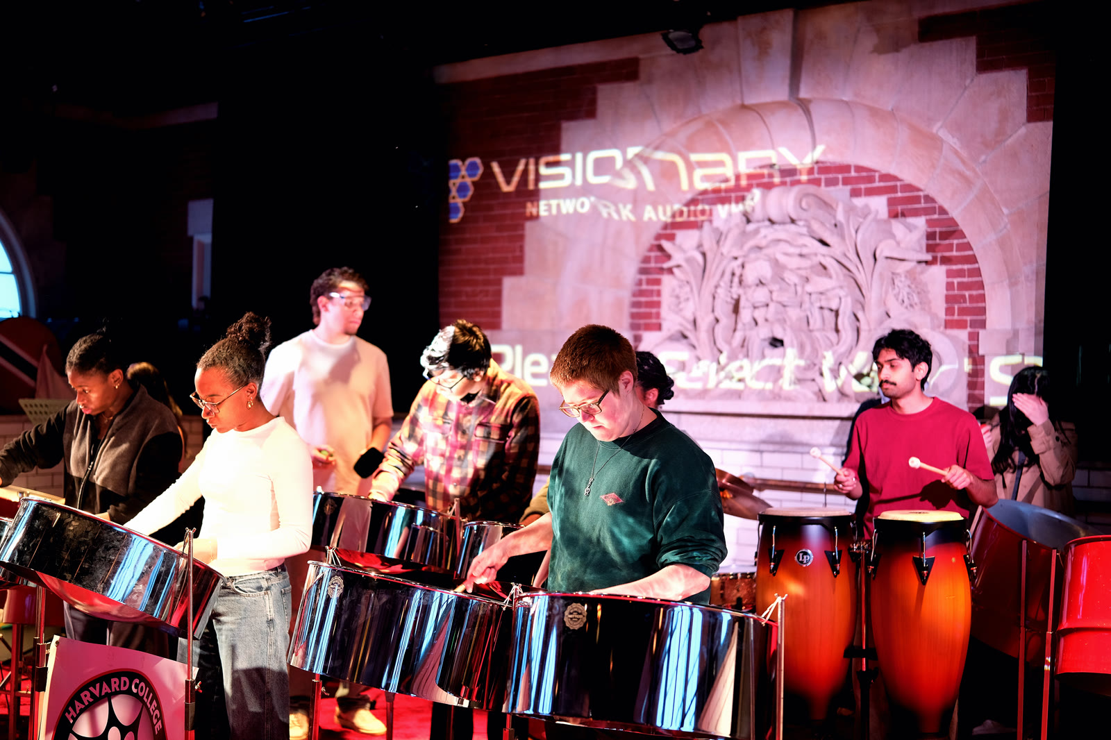 The ensemble rehearsing together on steelpans under colorful stage lighting.