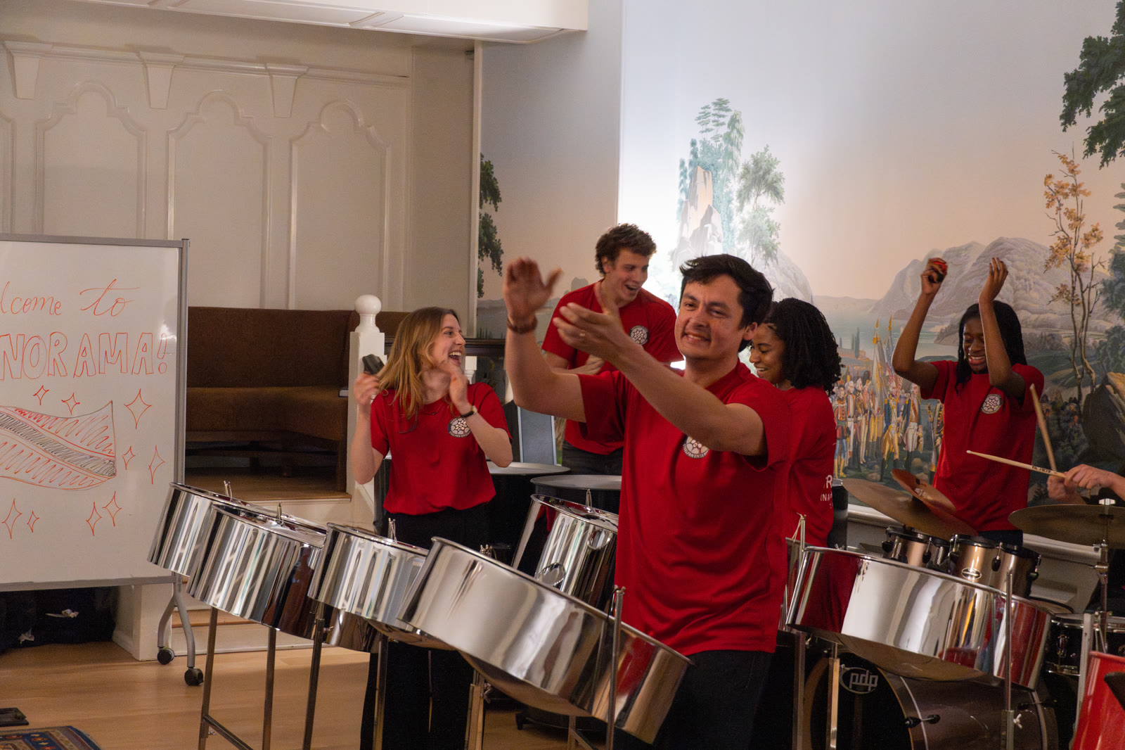 Ensemble members in matching red shirts smiling and playing steelpans during a rehearsal at Harvard.