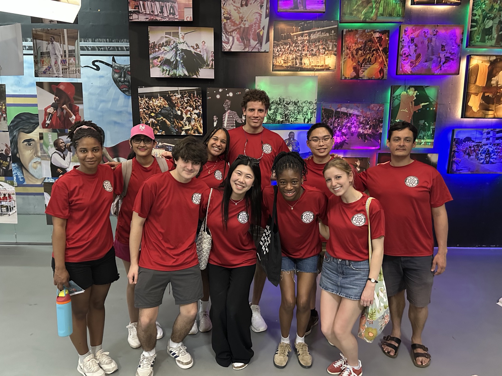 Ensemble members in red shirts in front of a wall covered in photographs of Carnival.
